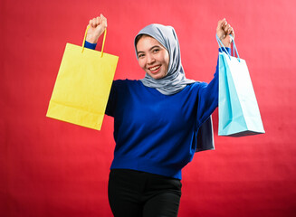 Asian woman wearing a gray hijab and blue sweater holds up yellow and blue shopping bags with a...