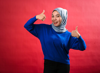 An Asian woman wearing a gray hijab and a blue sweater smiles while giving two enthusiastic...