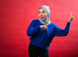 An Asian woman wearing a grey hijab and blue sweater points enthusiastically in two directions with...