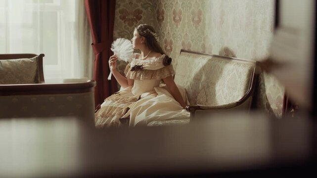 Beautiful young noblewoman in elegant 19th century attire sitting with fan in hand on upholstered sofa by window in sunlit well-furnished room with classical decor elements