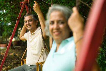 Mid age Indian couple with grey hair sitting on swings in a park, smiling, enjoying the sunny day