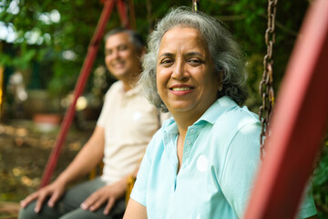 Obraz premium Mid age Indian couple with grey hair sitting on swings in a park, smiling, enjoying the sunny day