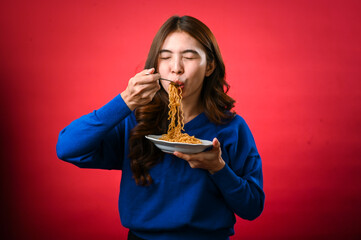 An Asian woman in a blue sweater happily slurps noodles from a plate, her eyes closed in enjoyment. She holds a fork and stands against a vibrant red background, savoring the delicious meal.