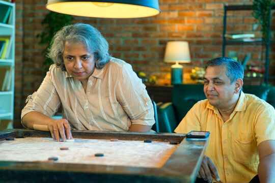Indian couple in their fifties playing carrom indoors, smiling, enjoying a fun and relaxed moment