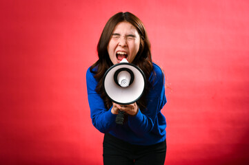 Fototapeta premium An Asian woman wearing a blue sweater is holding a red and white megaphone while speaking into it. She appears to be shouting or making an announcement. The background is solid red