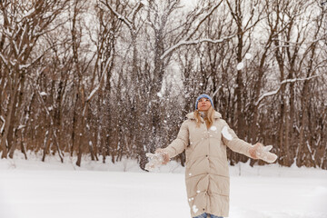 Naklejka premium Winter lifestyle portrait of cheerful pretty girl in stylish knitted hat throwing snow in the air. Smiling and having fun in the snow park. Snowflakes falling down. Freedom and happiness concept.