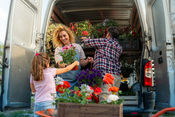 A family of three loading colorful flowers from a wheelbarrow into a delivery van for transport,...