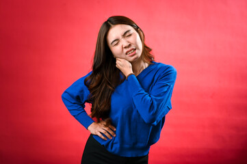 An Asian woman in a blue sweater, with long brown hair, is grimacing in pain as she holds her neck...