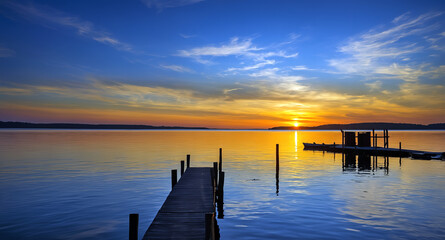 Obraz premium Perspective view of wooden pier over calm lake at sunset with dramatic sky