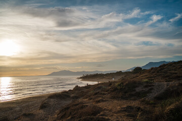 The golden light of sunset bathes the coastal dunes as distant mountains provide a stunning backdrop. The peaceful shoreline adds to the tranquil and scenic view