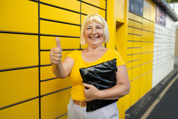 Senior 50s woman shows like getting parcel from cell of automatic post terminal outdoors. Concept of contactless and smart delivery. Idea of modern shipping and logistics. Woman wearing yellow t-shirt © Shi 