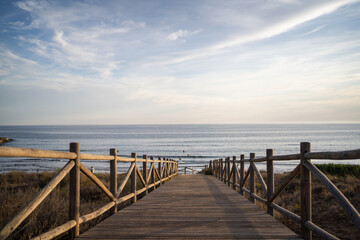 Obraz premium A wide-angle view of a peaceful beach and ocean as the sun sets behind distant hills, casting a warm glow over the coastline and sea