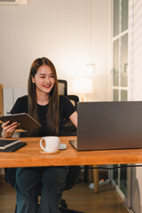 young woman is sitting at wooden desk, smiling while using laptop and holding tablet. modern office setting features warm lighting and cozy atmosphere, perfect for productivity