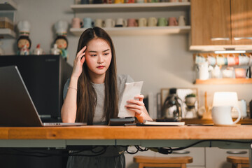 young woman is sitting at desk, looking concerned while reviewing document. setting is cozy kitchen with various mugs displayed on shelves, creating warm atmosphere