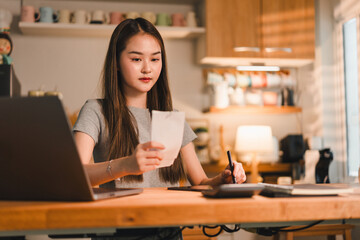 young woman is focused on her work at wooden desk, holding piece of paper while using laptop and tablet. cozy workspace is well lit, creating productive atmosphere