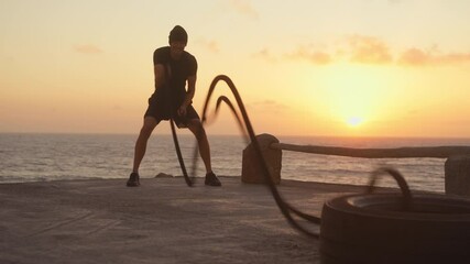 Determined sporty man in activewear training with battle ropes on platform by sea during sunset, casting beautiful backdrop to his intense workout 