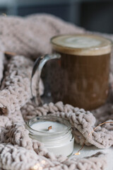 A cozy still life. A candle with a wooden wick and black coffee with milk in a glass mug on the table in a beige knitted plaid. Place for the text