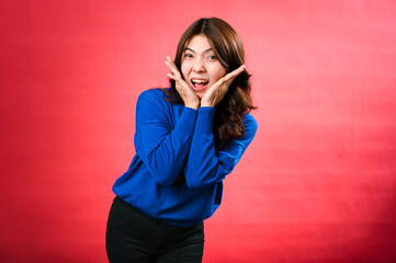 An Asian woman in a blue sweater expresses happiness with a big smile, her hands framing her face in a playful manner. She stands against a bright red background, radiating joy and positivity