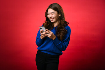 An Asian woman wearing a blue sweater is smiling as she looks down at her phone, engrossed in...