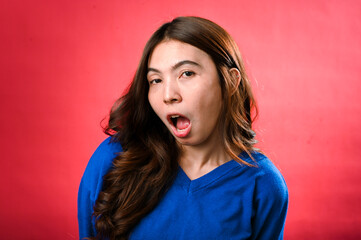 A young Asian woman with long, wavy brown hair poses confidently against a vibrant red backdrop. She is wearing a bright blue sweater and black pants, smiling softly with a casual stance