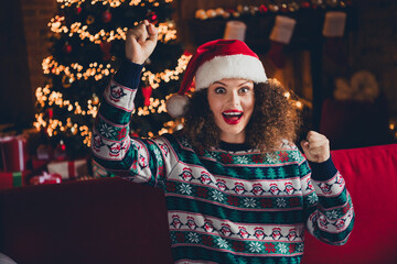 Portrait of nice young woman sit sofa raise fists wear sweater new year xmas garland lights flat indoors