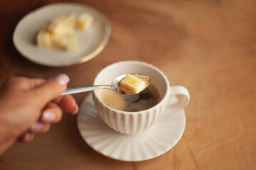 Butter coffee or bulletproof coffee, a high-calorie, high-fat drink free from carbohydrates to start the day; a woman's hand adding a spoonful of butter to a cup of coffee