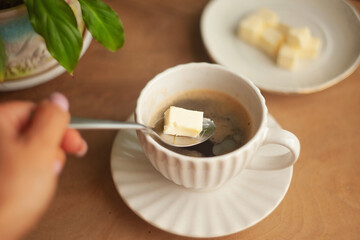 Butter coffee or bulletproof coffee, a high-calorie, high-fat drink free from carbohydrates to start the day; a woman's hand adding a spoonful of butter to a cup of coffee