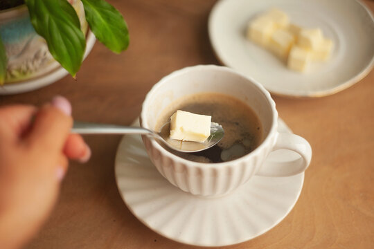 Butter coffee or bulletproof coffee, a high-calorie, high-fat drink free from carbohydrates to start the day; a woman's hand adding a spoonful of butter to a cup of coffee