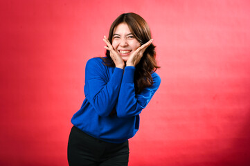 An Asian woman in a blue sweater expresses happiness with a big smile, her hands framing her face in a playful manner. She stands against a bright red background, radiating joy and positivity