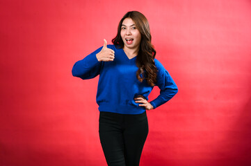 An Asian woman in a blue sweater and black pants stands confidently against a red background. She...