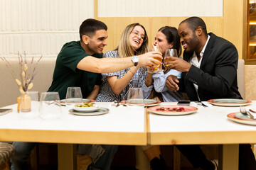 A group of 4 young people of different ethnicity between 20 and 30 years old are happily toasting with beer at the table of a modern restaurant. Concept of celebrating events.