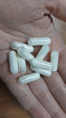 a hand holding several white capsules. The capsules are cylindrical and appear to be filled with a powdery substance. There are about nine capsules visible in the hand