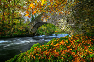 Lake District autumn colours at Clappersgate packhorse bridge with Brathay Holy Trinity Church in background.