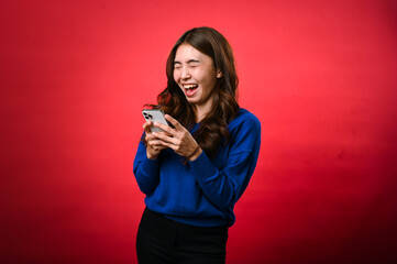 An Asian woman wearing a blue sweater is smiling as she looks down at her phone, engrossed in...