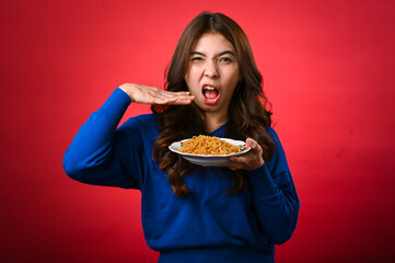 An Asian woman wearing a blue sweater holds a plate of noodles in one hand while gesturing excitement with the other hand near her mouth. She stands against a red background