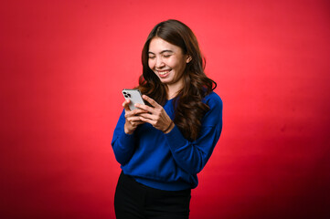 An Asian woman wearing a blue sweater is smiling as she looks down at her phone, engrossed in typing or reading a message. She stands against a bright red background in a cheerful, casual pose