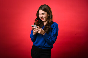 An Asian woman wearing a blue sweater is smiling as she looks down at her phone, engrossed in typing or reading a message. She stands against a bright red background in a cheerful, casual pose