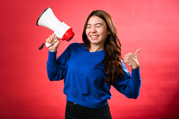 An Asian woman wearing a blue sweater is holding a red and white megaphone while speaking into it. She appears to be shouting or making an announcement. The background is solid red