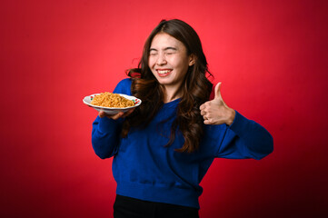 An Asian woman wearing a blue sweater smiles enthusiastically while holding a plate of noodles in one hand and giving a thumbs-up with the other. She stands against a bright red background