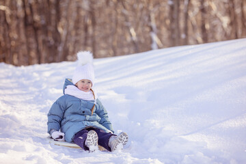 Happy little girl in warm blue jacket and white knitted hat and scarf riding down a snow slide in...
