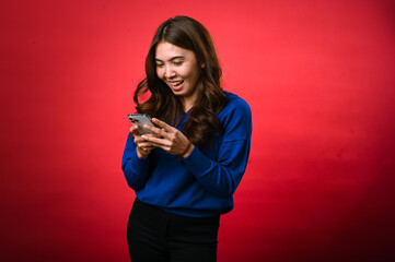 An Asian woman wearing a blue sweater is smiling as she looks down at her phone, engrossed in...