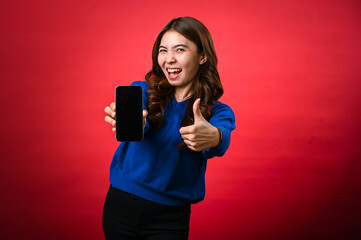 An Asian woman in a blue sweater holds a smartphone with a black screen in one hand and gives a thumbs-up with the other. She smiles confidently against a vibrant red background.