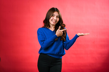 Fototapeta premium An Asian woman in a blue sweater presents with one hand outstretched and the other pointing towards it. She smiles confidently, standing against a bright red background in a casual, friendly pose