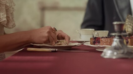 Cropped shot of unrecognizable 19th century woman in elegant dress spreading butter on bread while having tea party with noble people in wealthy dining room with classical decor