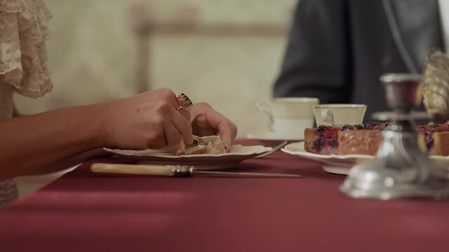 Cropped shot of unrecognizable 19th century woman in elegant dress spreading butter on bread while having tea party with noble people in wealthy dining room with classical decor