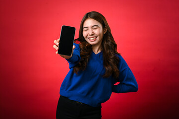 Naklejka premium An Asian woman wearing a blue sweater is smiling excitedly while holding a smartphone forward in her hand, displaying its screen to the viewer. The background is a bold, solid red