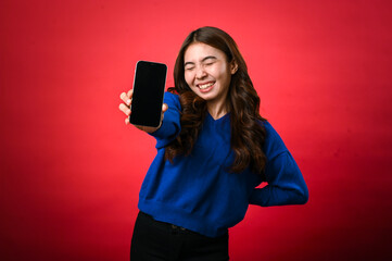 Naklejka premium An Asian woman wearing a blue sweater is smiling excitedly while holding a smartphone forward in her hand, displaying its screen to the viewer. The background is a bold, solid red