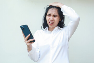 Naklejka premium portrait of frustrated asian young woman manager using smartphone touching her head, unbelieve dissatisfied face expression, annoyed female office worker employee wearing white formal shirt, standing