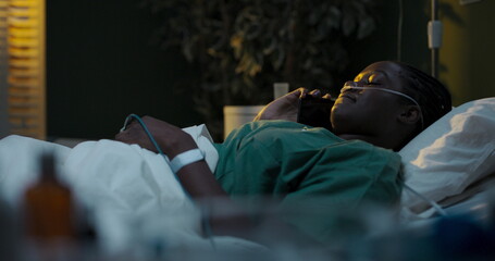 Young African patient in hospital with oxygen tube and oximeter talks on phone, tears visible.