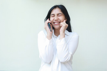 ecstatic beautiful asian business woman entrepreneur laughing with closed eye while speaking on mobile phone and raising palms wearing white formal suit shirt, celebrate winning victory, standing